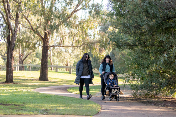 Two adults walk along a paved park path in Heathdale Ward's Riverbend Historical Park beside tall trees, with one adult pushing a stroller carrying a small child, in a green, sunlit outdoor setting.