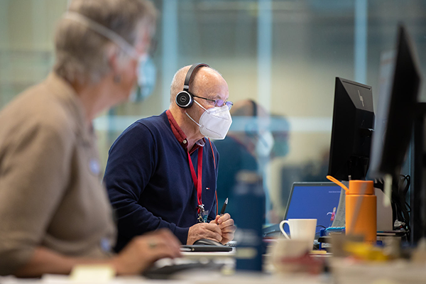 A man wearing a face mask and a telephone headset. He is sitting at a desk looking at a computer.