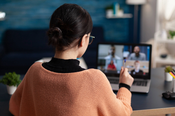 A person in an orange knit top and glasses faces a laptop screen with a meeting on the screen