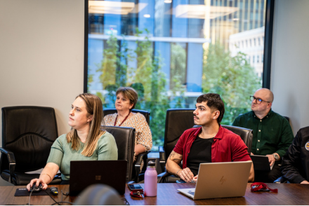 A modern office meeting room with 4 people watching a presentation. Two people in the foreground are using laptops. Behind them, 2 others are seated near large windows with views of trees and office buildings. 