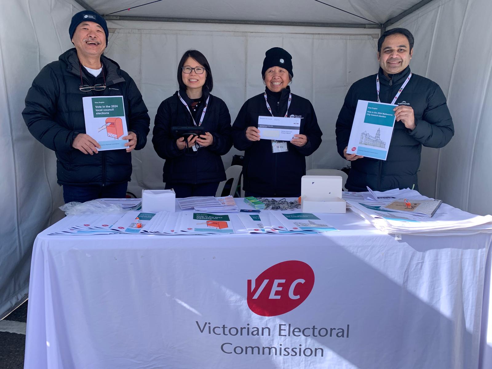 Four VEC staff members stand behind an information table at an electoral outreach session. They each hold various election information materials in their hands.