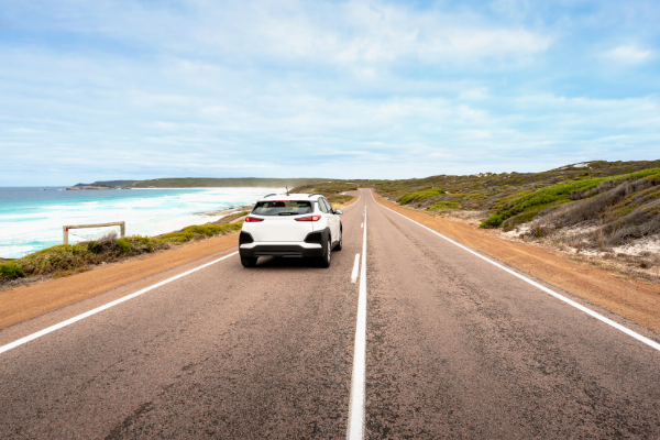 A white care driving on the left hand side of the road with the ocean on the left