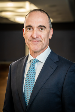 A professional portrait of the Victorian Electoral Commissioner, Sven Bluemmel, in a navy blue suit, white shirt, and orange plaid tie, standing against a plain grey background.