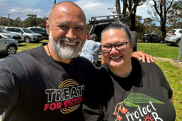 Two people smiling and posing for the camera. They are wearing a 'Treaty for Victoria' shirt and another shirt with a gum tree design.